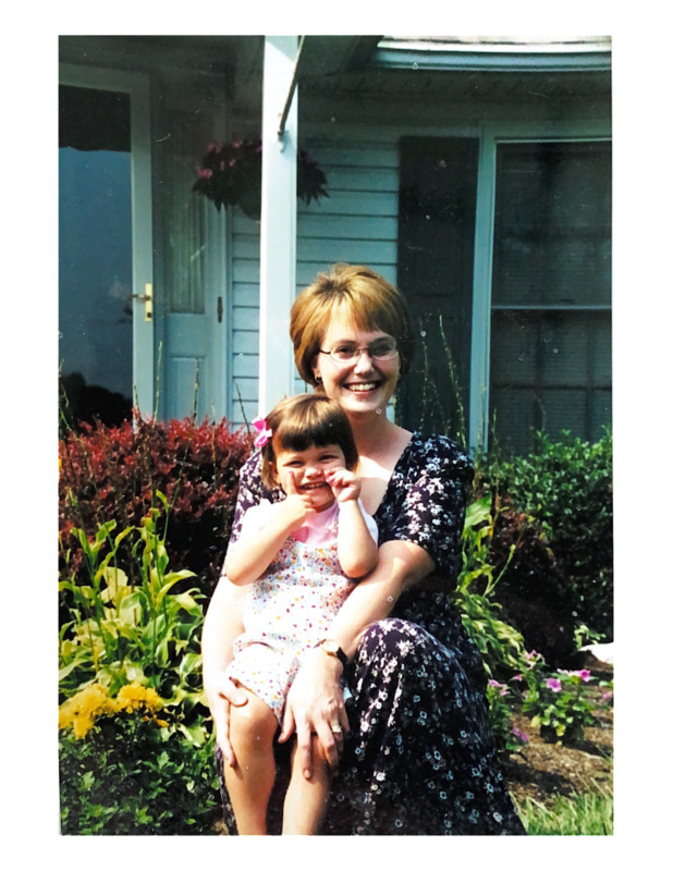 Scan of a color photograph of Lisa and Madison Cissell outside of their home in Versailles, Kentucky. There are sitting in front of their lawn, with lots of plants in the background. Lisa is wearing a floral dress and Madison is wearing floral-pattern overalls with a pink bow in her hair. 