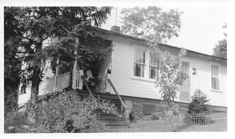 Black and white photograph of a small white house with a side, screened-in porch surrounded by trees and shrubs.