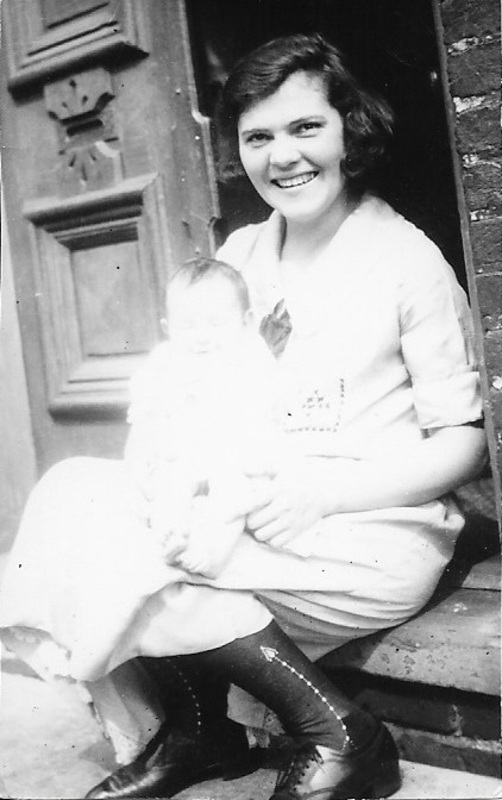 Black and white photograph of Mina Montgomery, a smiling white woman wearing a dress, black socks, and black shoes, sitting on a door stoop holding a baby in Cumberland, Maryland. The photograph is overexposed. 