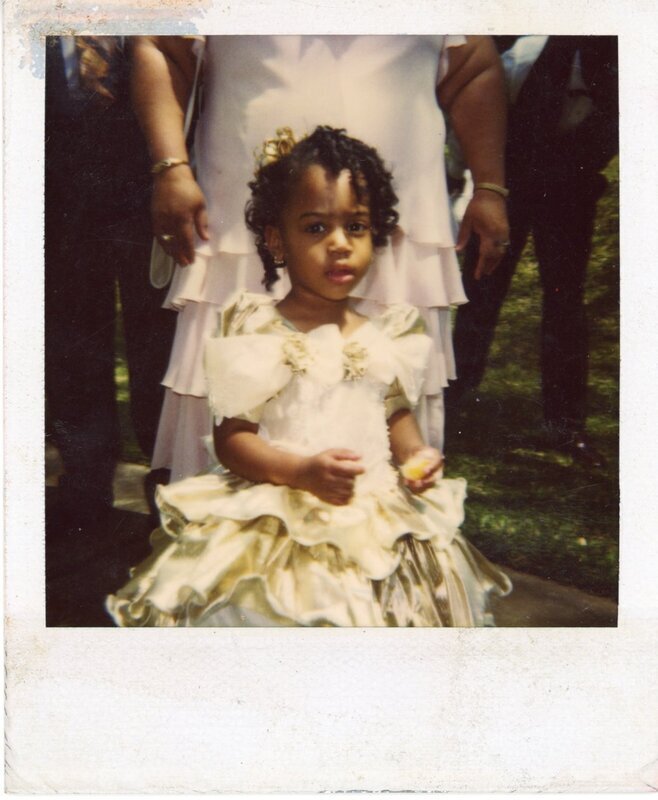 Scan of a color photograph of Sierra King in Gold Dress. Sierra in gold dress standing in front of Mamarita holding a piece of candy or cake looking at the camera 