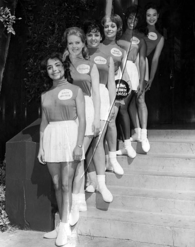 black and white photo of six young women, representing various nationalities, wearing matching outfits and posing in a line down a staircase. They are wearing nametags to represent a specific continent or region