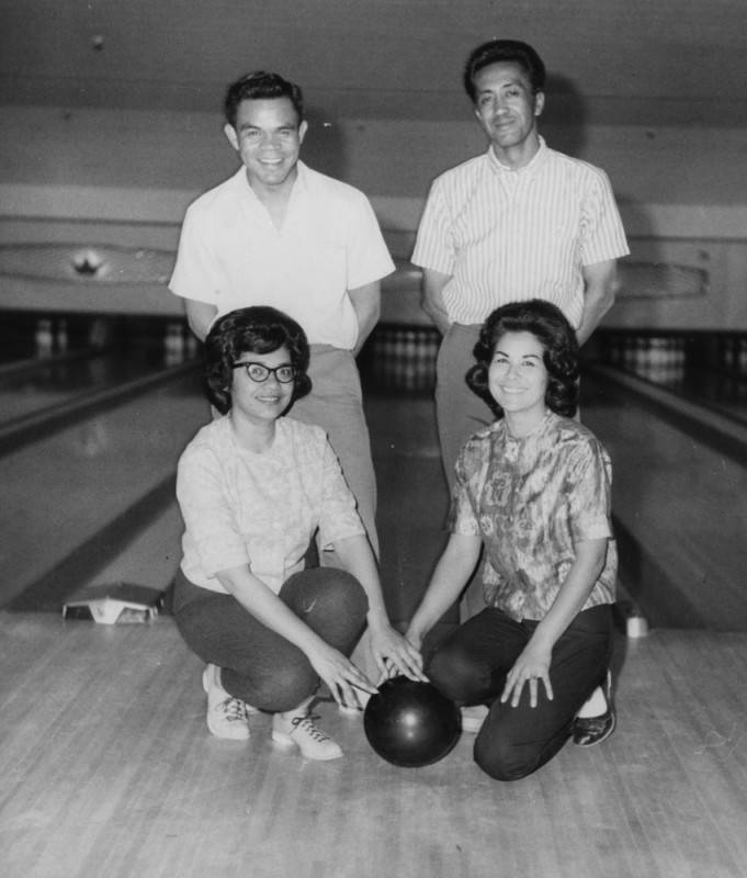 black and white photo of four bowlers at a bowling alley, two men standing and two women kneeling in front. The two women have hands placed on a bowling ball in between them.