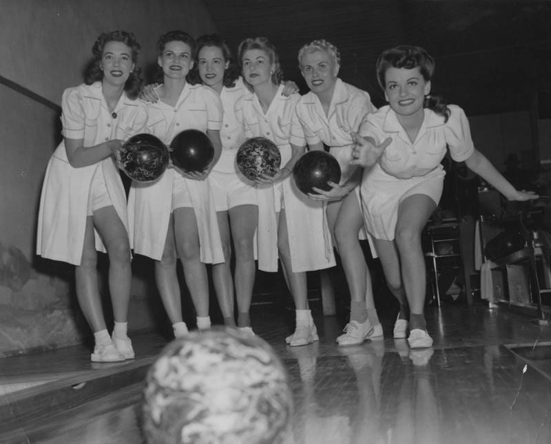 black and white photo of six young women wearing matching uniforms while standing on a bowling lane. One of the women has just rolled her bowling ball while the five other women hold their bowling balls and pose for the photo.