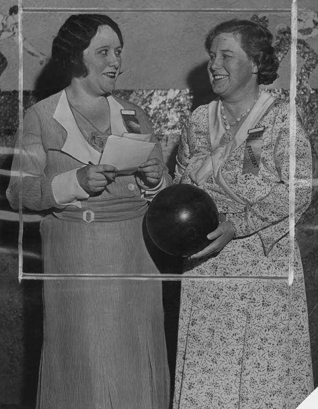 black and white photo of two women facing one another, smiling, while one holds a paper. They each have ribbons pinned to the front of their dresses, one visibly reading "President."