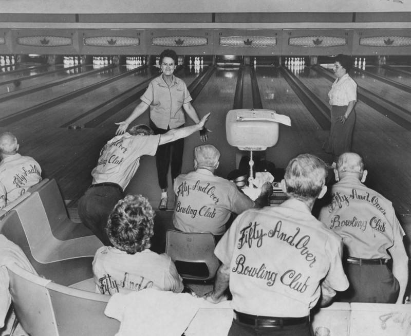 black and white photo of group of older bowlers at a bowling alley. They have on matching shirts with the phrase "Fifty And Over Bowling Club" printed or embroidered in script letters on the back of them. 