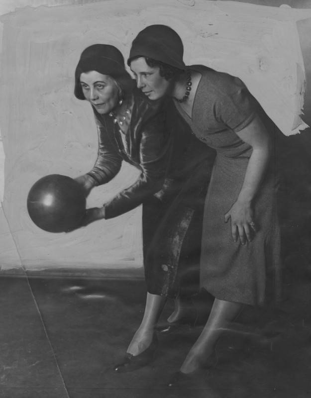 black and white photo of two women bowling at a bowling alley. One is holding a bowling ball, as if preparing to roll, and the other stands nearby. They are both wearing dresses, hats, and hoisery and one dress appears to be dark velvet.