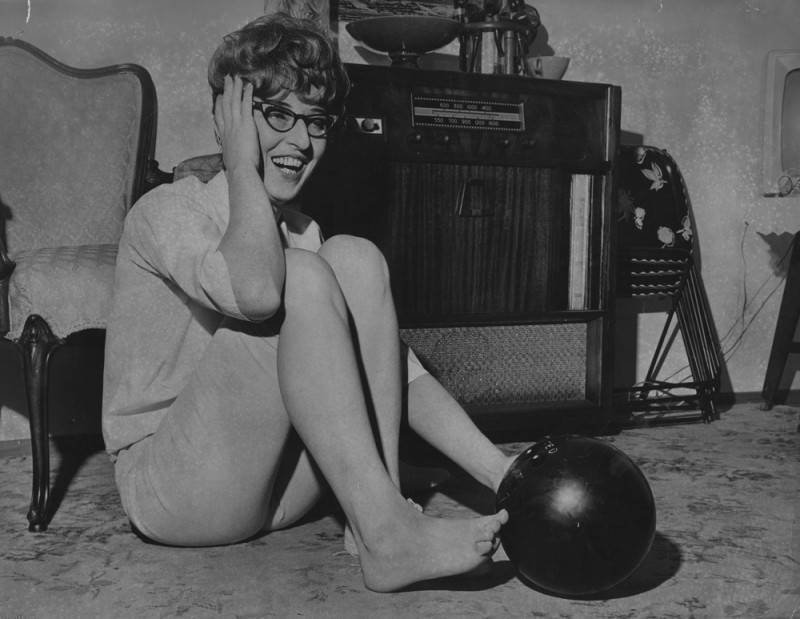 black and white photo of a young woman sitting on a rug in her home with the big toe of her right foot stuck inside of a bowling ball's finger holes. She is smiling and holding her hand to the side of her head. There is a radio, chair, and TV trays visible in the background.