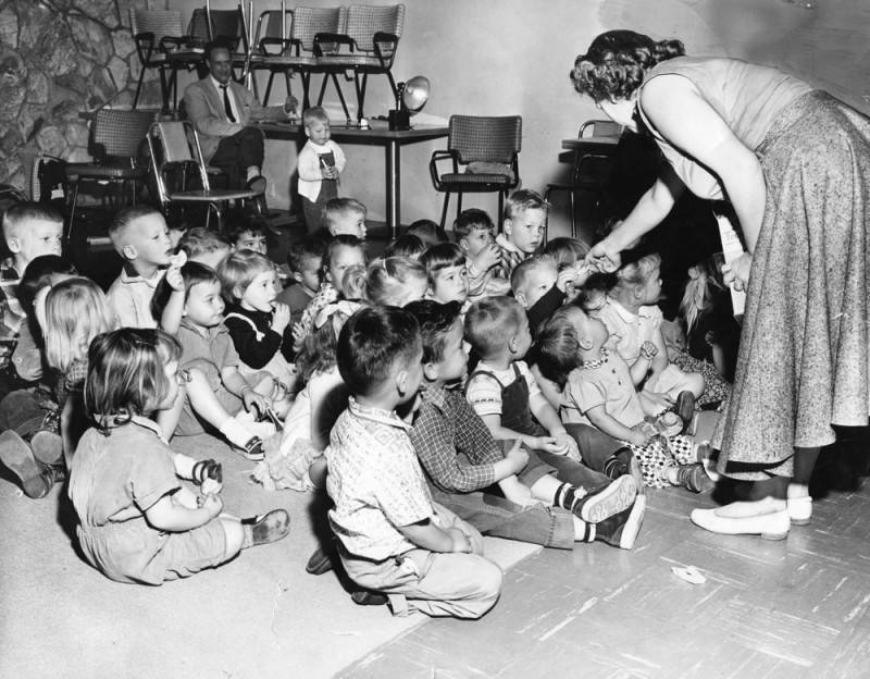 black and white photo of about two dozen young children sitting on a rug while a woman hands out snacks to them. There is a man sitting in the back of the room.