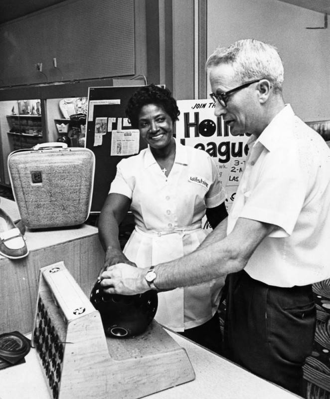 black and white photo of a smiling woman wearing a "Wilshire" uniform, smiling at a man as she tests a bowling ball for finger fit. 