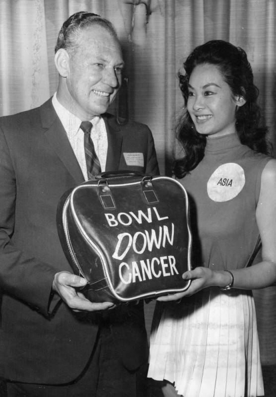 black and white photo of a businessman and an actress/model posing for a photo while holding a bowling ball bag with "Bowl Down Cancer" printed on its front in white letters.