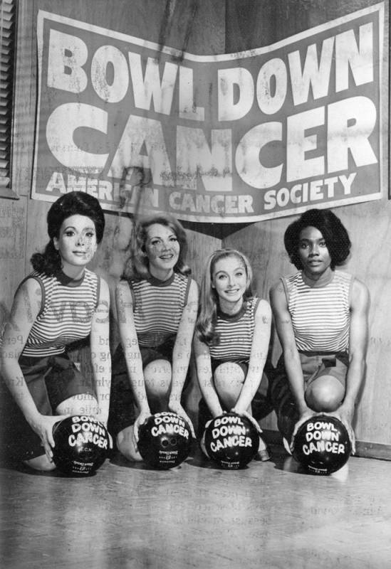 black and white photo of four actresses/models posing for a promotional photo. There is a banner behind the women with "Bowl Down Cancer" and "American Cancer Society" printed on it. The women are wearing matching outfits and each are holding a bowling ball with "Bowl Down Cancer" printed on it.