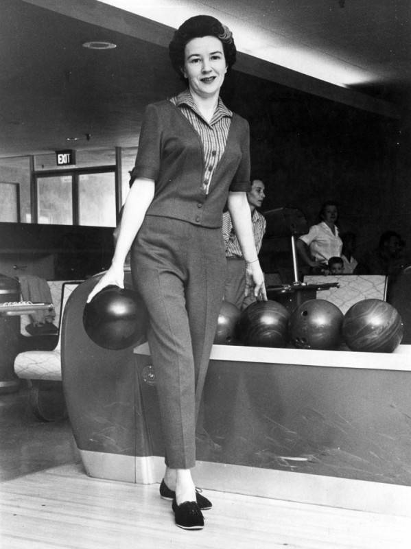 black and white photo of a woman bowler posing in front of a bowling ball retriever at a bowling alley. She is wearing a two-piece pantsuit set with prominent pleats in the trousers, along with a striped and collared shirt, and is holding a bowling ball in her hand.