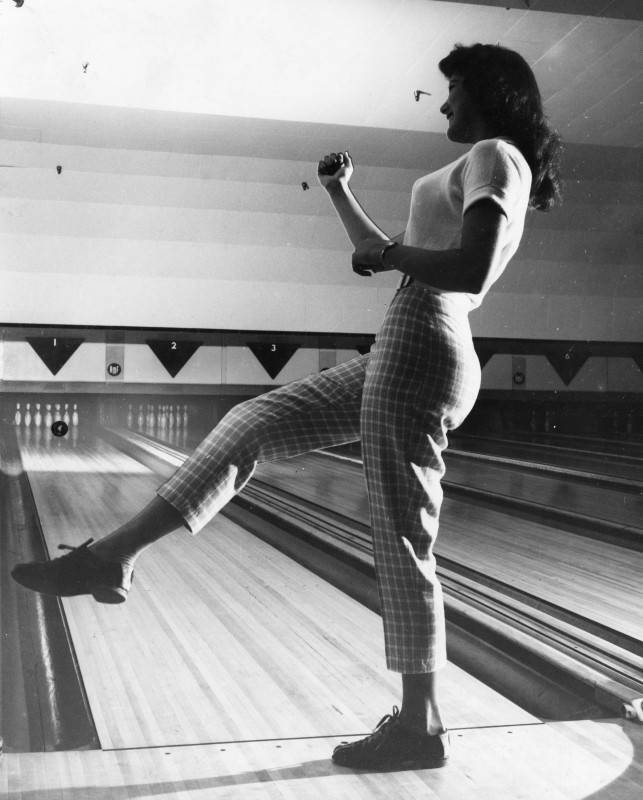 black and white side profile photo of a young woman bowler standing in front of a bowling lane with her arm and leg raised in a celebratory stance. 