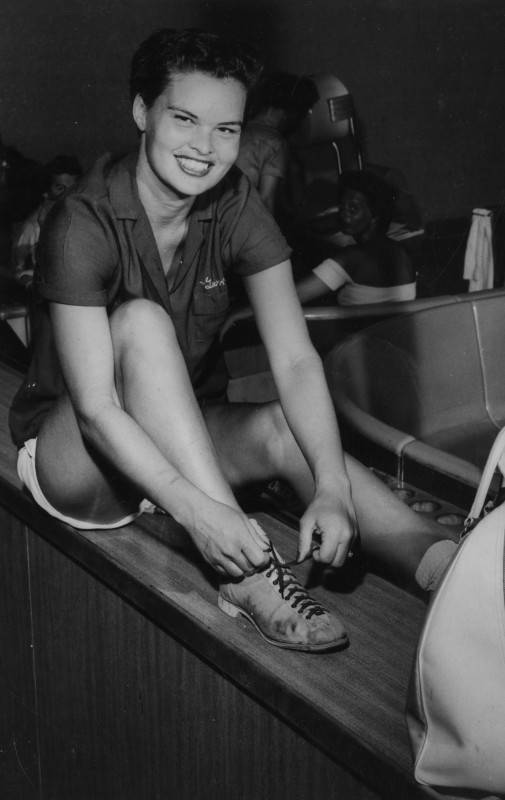 black and white photo of a young woman bowler sitting on a ledge in a bowling alley as she ties her bowling shoes and smiles at the camera.