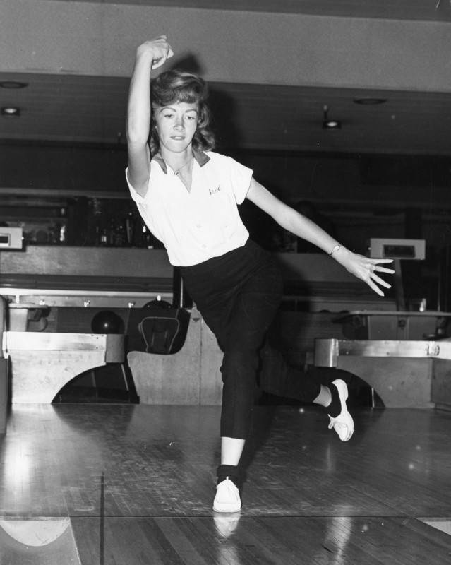 black and white photo of a young woman bowler at a bowling alley as she finishes a roll down the lane.