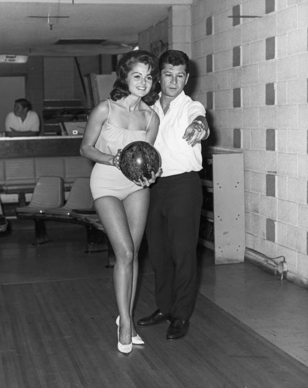 black and white photo of a man pointing down the lane of a bowling alley as a young woman in heels smiles while holding a bowling ball.