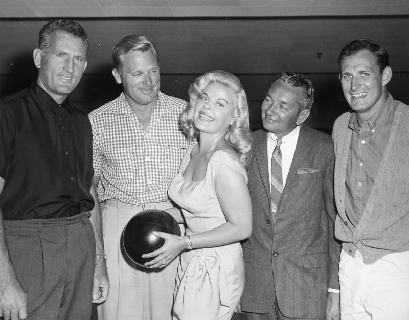 black and white photo of businessmen and an actress posing in a bowling alley for a promotional photo. The woman is smiling and posed in front of the group while holding a bowling ball in her hands. The four men are either smiling for the photo or looking at the actress.