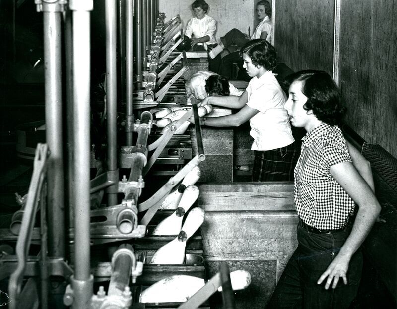 black and white photo of five young women resetting the pin machines, among other bowling-related tasks, in a bowling alley.