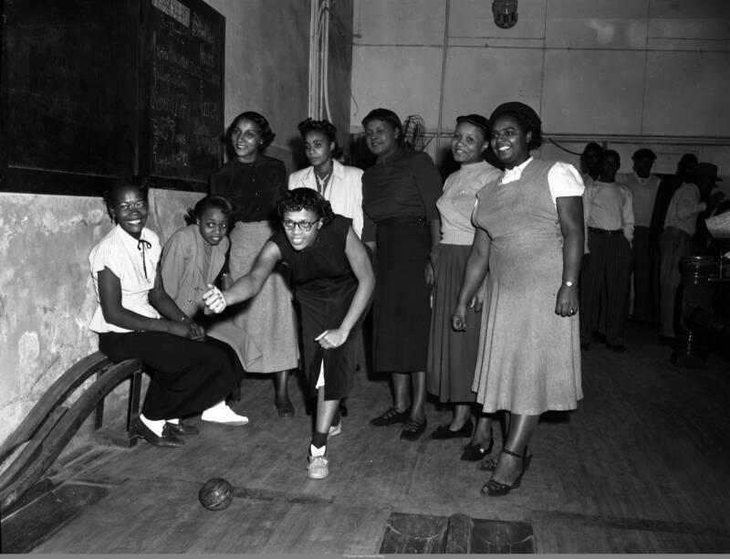 black and white photo of a group of African-American women bowlers bowling in a bowling alley with several onlookers behind them. One woman is posed as if she has just rolled her bowling ball while the other women are standing and smiling while looking in the direction of where the ball was rolled. Two women are sitting.