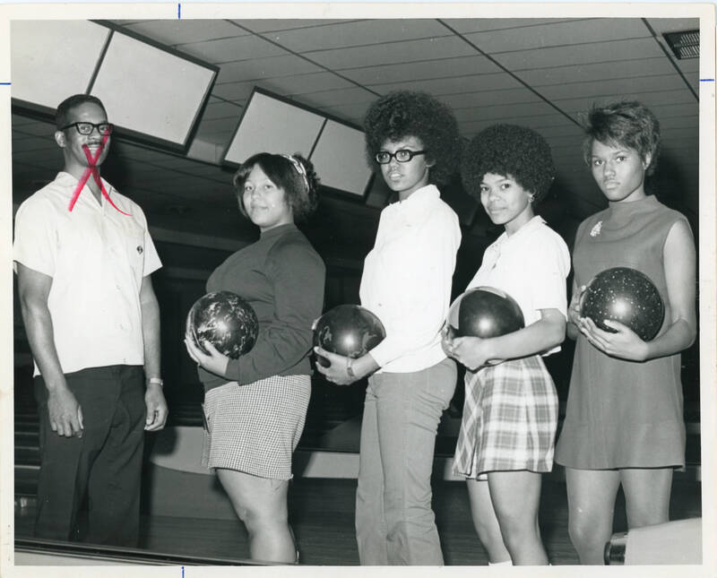 black and white photo of four African-American women bowlers posing inside of a bowling alley. There is also an African-American man in the photo with a red "x" marked over his face. The women are each holding a bowling ball while standing in a line.