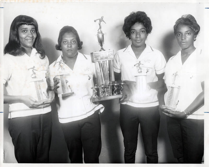 black and white photo of four African-American women bowlers in matching uniforms posing with a group bowling trophy and four individual trophies. Each woman is wearing a collared white shirt with their names embroidered above the chest pocket and black pants. 