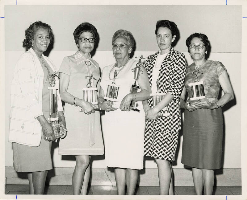 black and white photo of five older African-American women bowlers posing with their bowling trophies. One woman in the center is holding two trophies, the larger one presumably their group trophy.