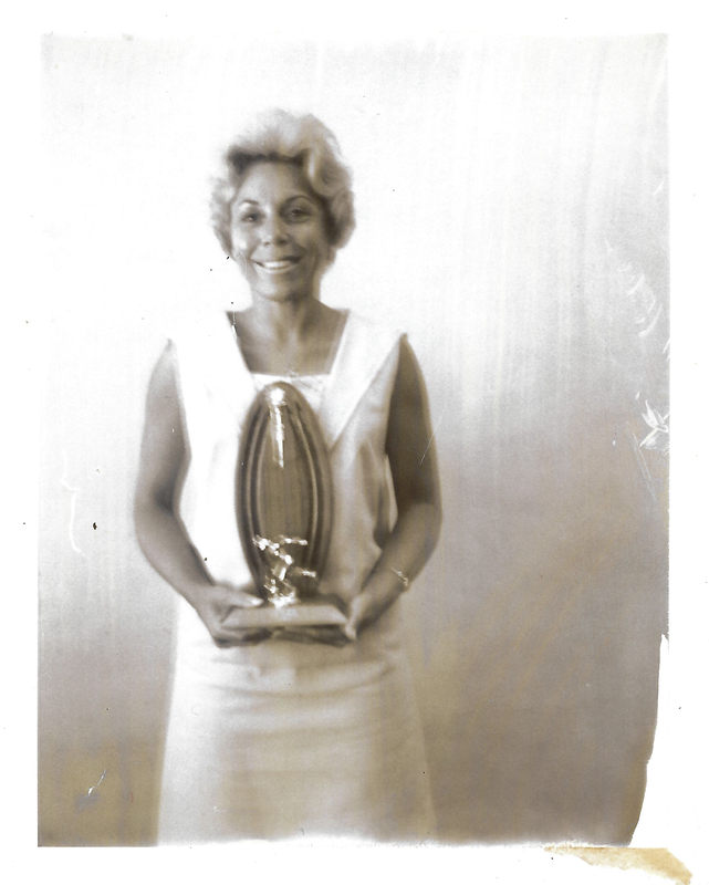 black and white, almost sepia-toned, photo of Bettye Dean smiling while holding a bowling trophy. She is wearing a sleeveless, light-colored dress and her hair is styled in short blonde waves.