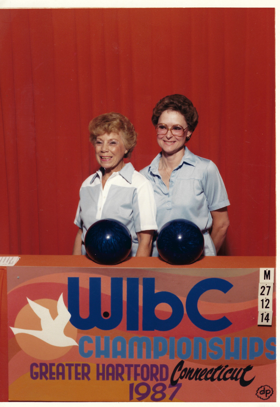 color photo of Bettye Dean and an unnamed team member posing for a portrait at the 1987 WIBC Chamipionships competition. The two women are standing behind an orange podium wearing a light blue uniform with slight variations between them, each with a blue bowling ball in front of them. The orange podium has "WIBC Championships Greater Hartford Connecticut 1987" painted on it along with a white bird.