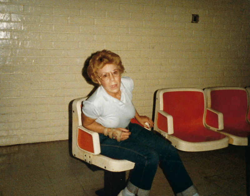 color photo of Bettye Dean sitting in a row of seats at a bowling alley. She is wearing glasses, a light blue collared shirt, denim pants with the hems cuffed, and a tan wrist/hand brace.