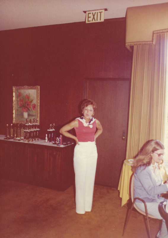 color photo of Bettye Dean posed in front of a long table with rows of bowling trophies lined up on it. She is smiling and wearing a sleevless red top, white pants, and a multi-colored white scarf around her shoulders.