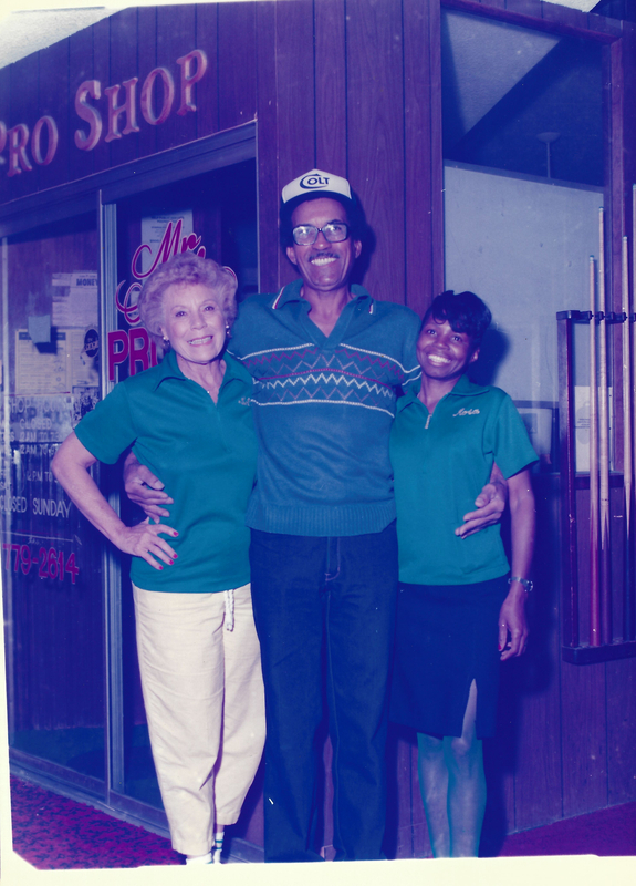 color photo of Bettye Dean posed with a man and woman in front of a bowling pro shop store window. They are all wearing dark green shirts with Bettye and the other woman wearing identical shirts with their names embroidered on the upper left chest area.