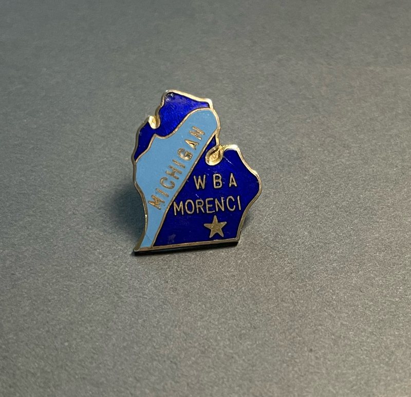 image of a lapel pin in the shape of the state of Michigan. The pin has dark and light blue enamel in the background and "Michigan WBA Morenci" and a star are inlaid in gold metal.