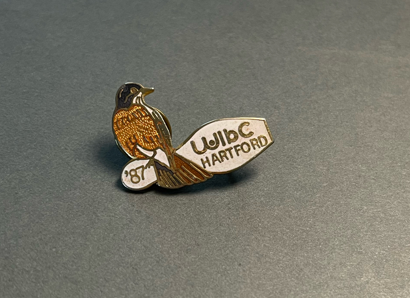 image of a lapel pin with an enamel and metal design of an American Robin perched atop a white bowling pin. On the bowling pin, "'87 WIBC Hartford" are inlaid in gold metal. 