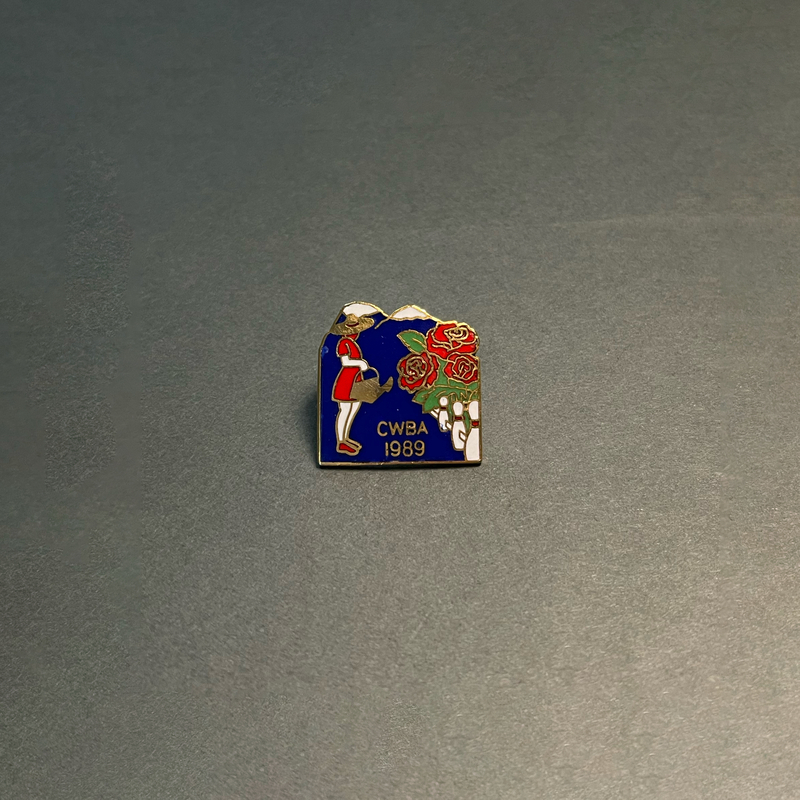 image of a square lapel pin with the shape of mountains at the top. The pin is an enamel and metal design with a blue background, white-capped mountains, and a woman in a red dress watering red roses behind a white fence made of bowling pins. "CWBA 1989" is inlaid in gold metal.