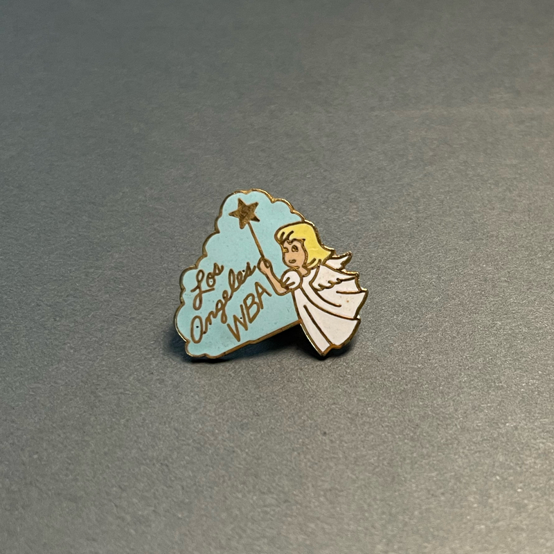 image of a lapel pin with an enamel and metal design of an angel amidst a light blue cloud. The angel has blonde hair and wings, and is wearing a white gown. "Los Angeles WBA" and a star-tipped wand are inlaid in gold metal.