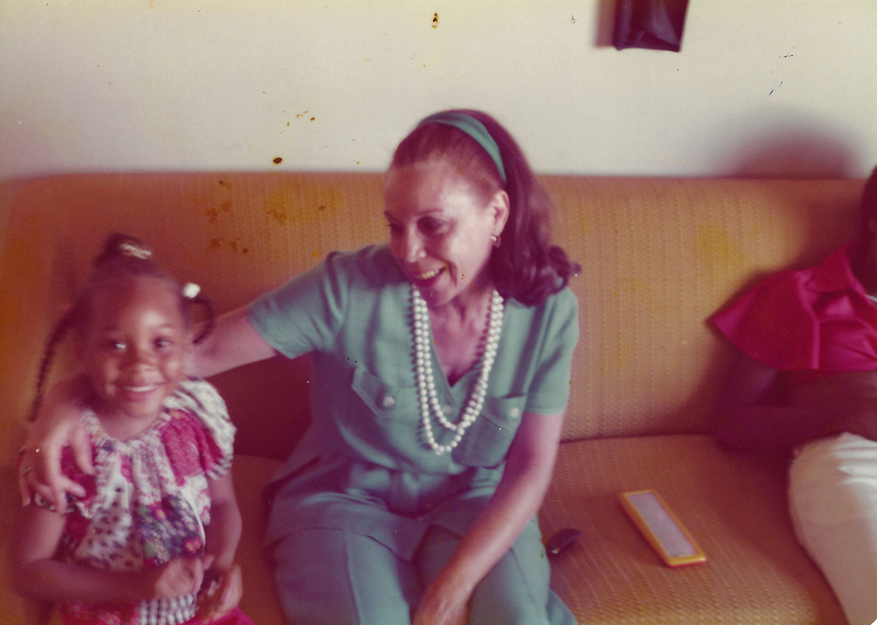 color photo of Nikki (White) Watkins as a child and her grandmother, Bettye (White) Dean sitting on a yellow couch, Nikki smiling at the camera and Bettye smiling down at her. Bettye is wearing a green pantsuit with a meatching green headband in her hair. Nikki is wearing a patchwork dress and her hair is in three braids decorated with hair balls and barrettes.