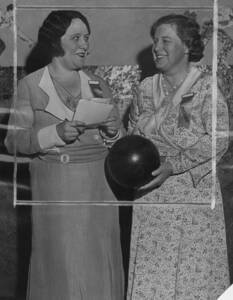 black and white photo of two women facing one another, smiling, while one holds a paper. They each have ribbons pinned to the front of their dresses, one visibly reading "President."