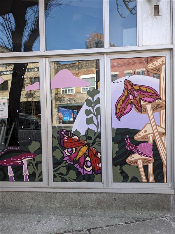 A store window, which has been painted to feature plants, butterflies, and mushrooms. A snail sits on top of one of the mushrooms. Shades of purple, pink, brown, and cream can be seen.