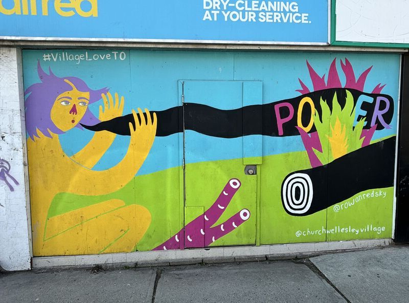 A painted mural on an old storefront featuring a yellow naked woman with purple hair blowing the word "power" onto multi-coloured flames