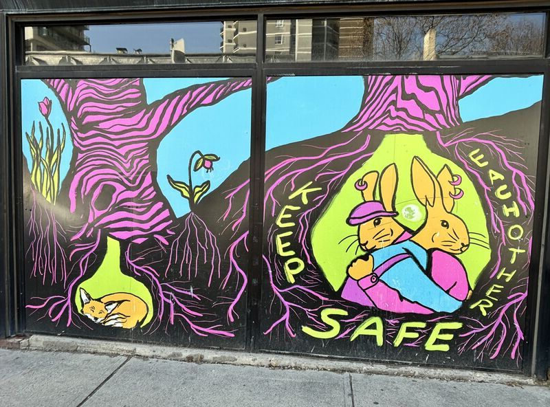 A mural painted on two windows featuring two purple trees, a sleeping fox, ans two clothed anthropmorphic rabbits hugging