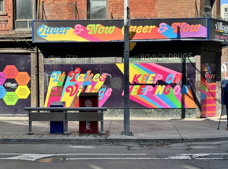 A brightly printed mural featuring the phrases "Queer & Now" twice, "It Takes a Village," "Keep On Keeping On," and "Take Care"