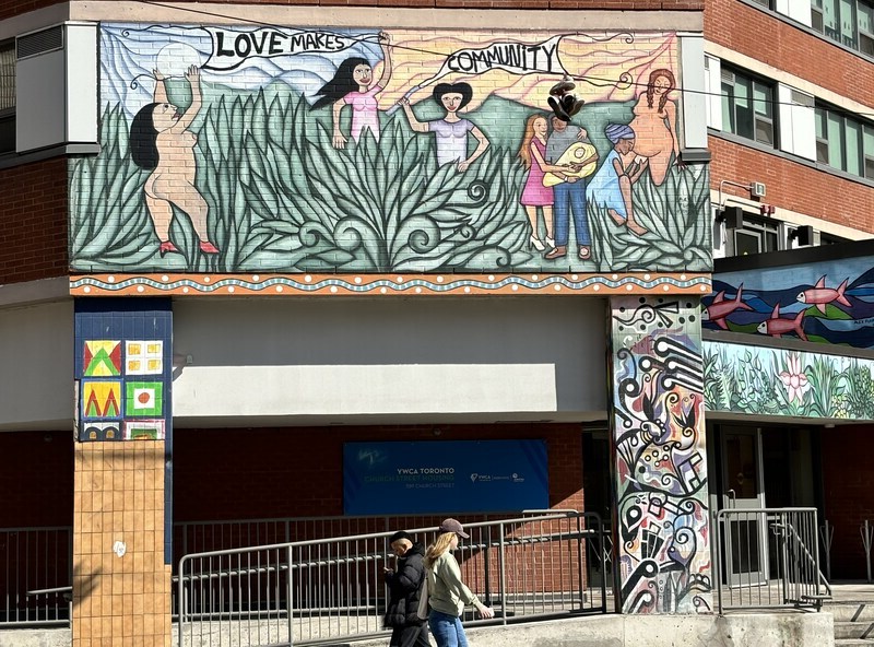 A mural painted on the side of a building featuring tall green grass, the moon and individuals doing a range of activities including giving birth, holding a baby, and holding up pennants with the phrase "Love Makes" and "Community"