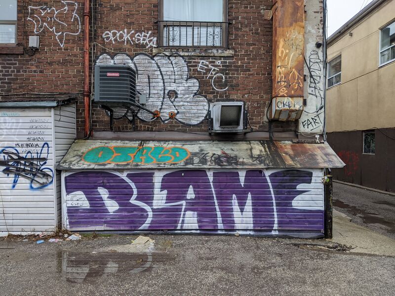 Tags on garbage bins in a back alley. BLAME is written in large, purple letters.