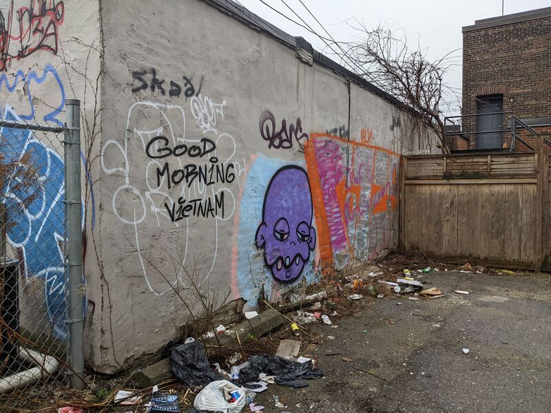 Tags on a wall near garbage in an alley, including "Good Morning Vietnam" and a purple head.