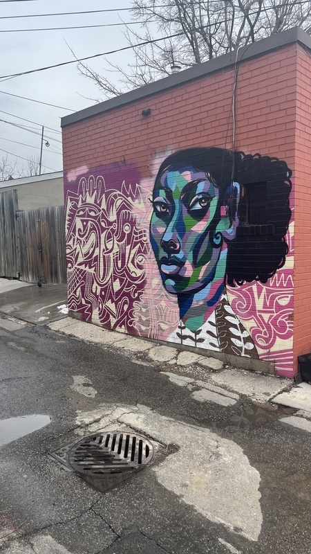 Painting of a woman painted with green, blue, pink, and purple on top of a geometric pattern on a brick wall.