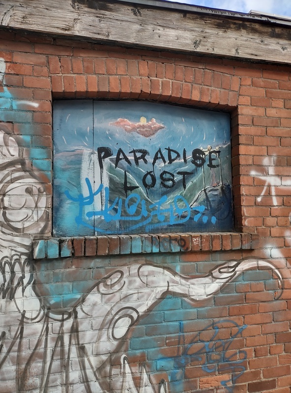 "PARADISE LOST" painted over a landscape on a boarded up hole in a brick wall surrounded by other tags. 