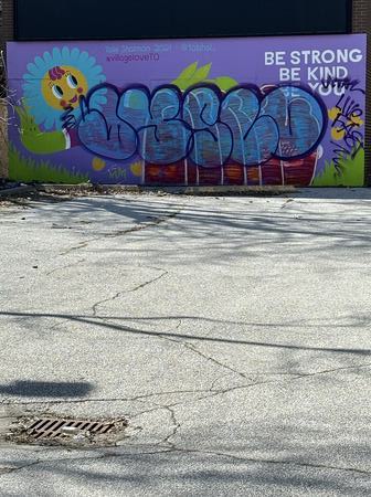 A mural featuring a smile face flower with the phrase "be strong, be kind, be you" on the front of an old boarded up store. Another graffiti tag has been painted over top and features the phrase "live fast"