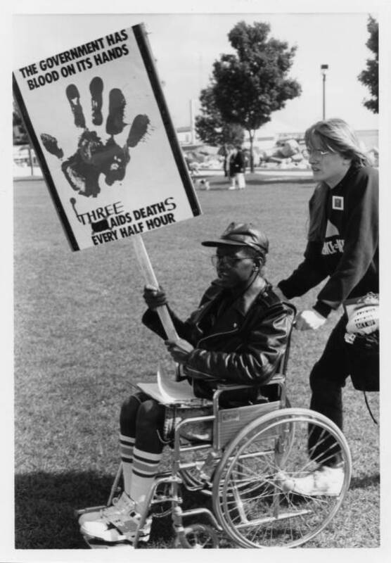 grayscale photograph depicting an ACT UP activist, assisting another ACT UP member in a wheelchair carrying protest sign. The sign reads "THE GOVERNMENT HAS BLOOD ON ITS HANDS... THREE AIDS DEATHS EVERY HOUR." A large splatter of a handprint is at the focal point of the sign.