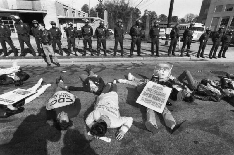 grayscale photograph of policemen lined-up standing in front of the Centers for Disease Control as activists from ACT UP stage a 'die-in' holding protest signs.