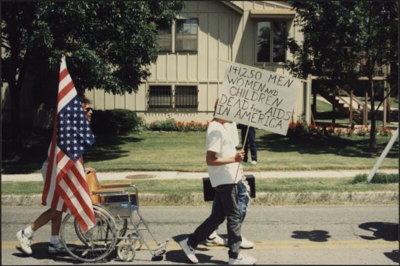 color photograph of man marching at a pride parade carrying a sign covering his face that reads "141,250 men, women and children dead from AIDS in America"; he is followed by another man pushing an empty wheel chair and carrying an American flag; a house they're passing is seen behind them.